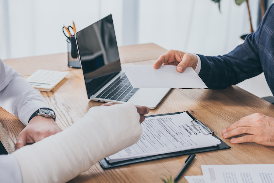 cropped view of businessman sitting at table and giving paper sheet to worker with broken arm in office, compensation concept what is workers compensation and how does it work